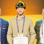 Five men in business attire stand together at a precast concrete event, one holding a certificate and wearing a construction hat.